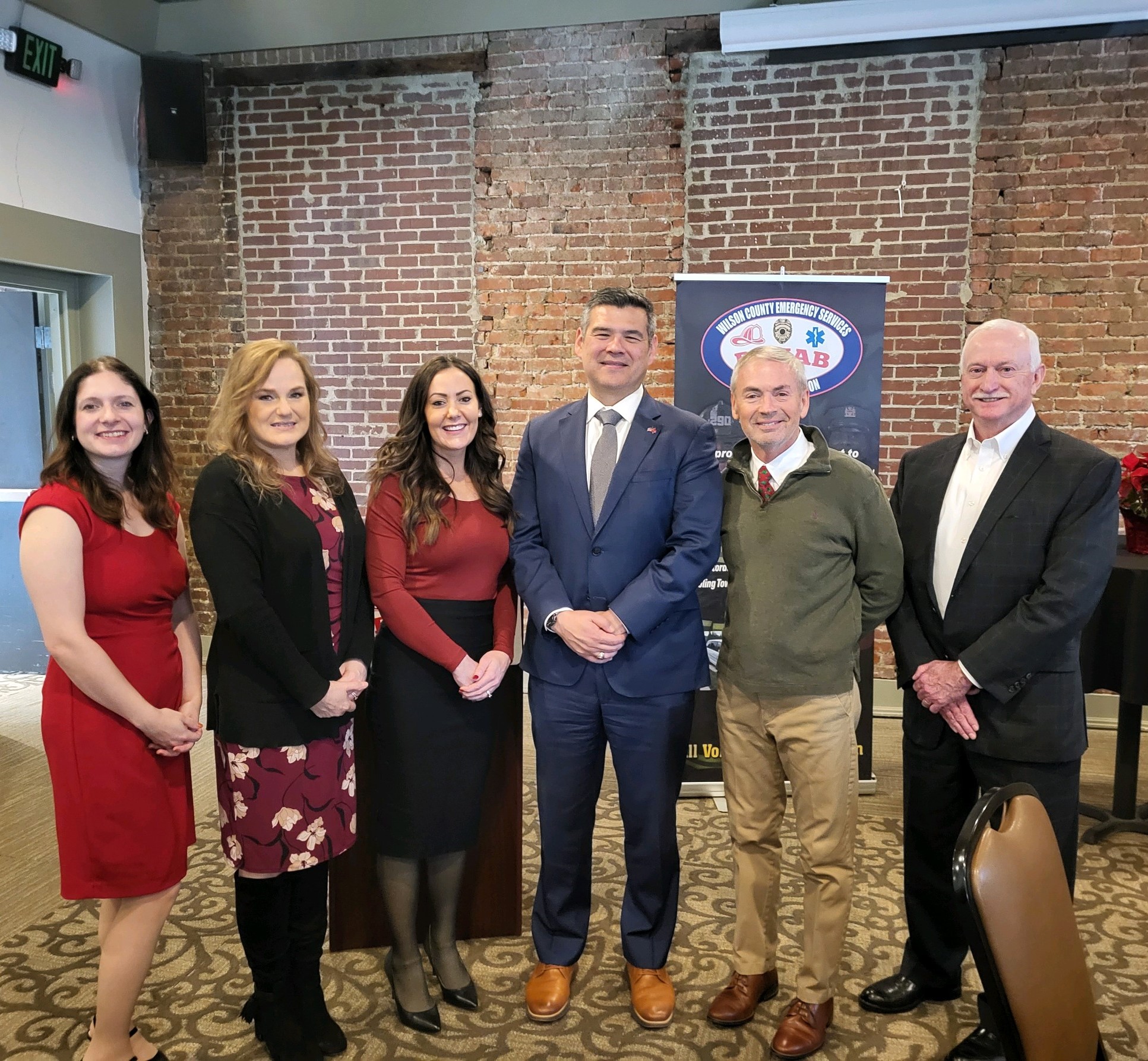 FFB Staff pose with guest speaker and mayor in front of a brick wall and a Rehab23 sign.